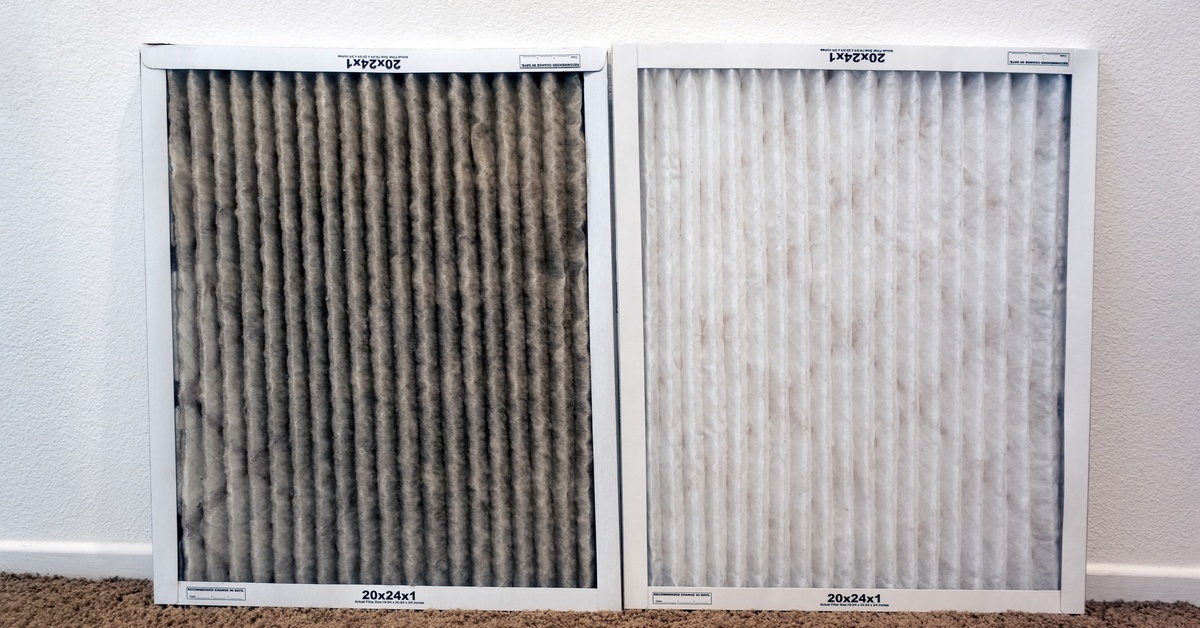 A dirty HVAC filter sits next to a clean HVAC filter. The filters lean against a white wall in a carpeted room.