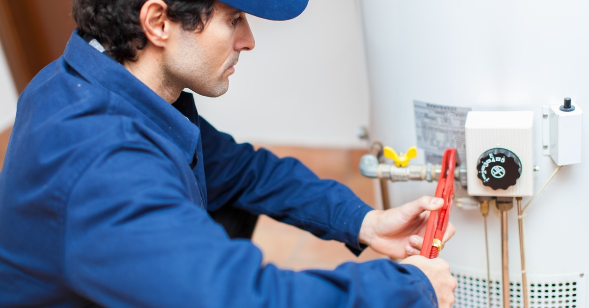 A plumber wearing a blue jumpsuit and baseball cap uses a wrench to make adjustments on a hot water heater.