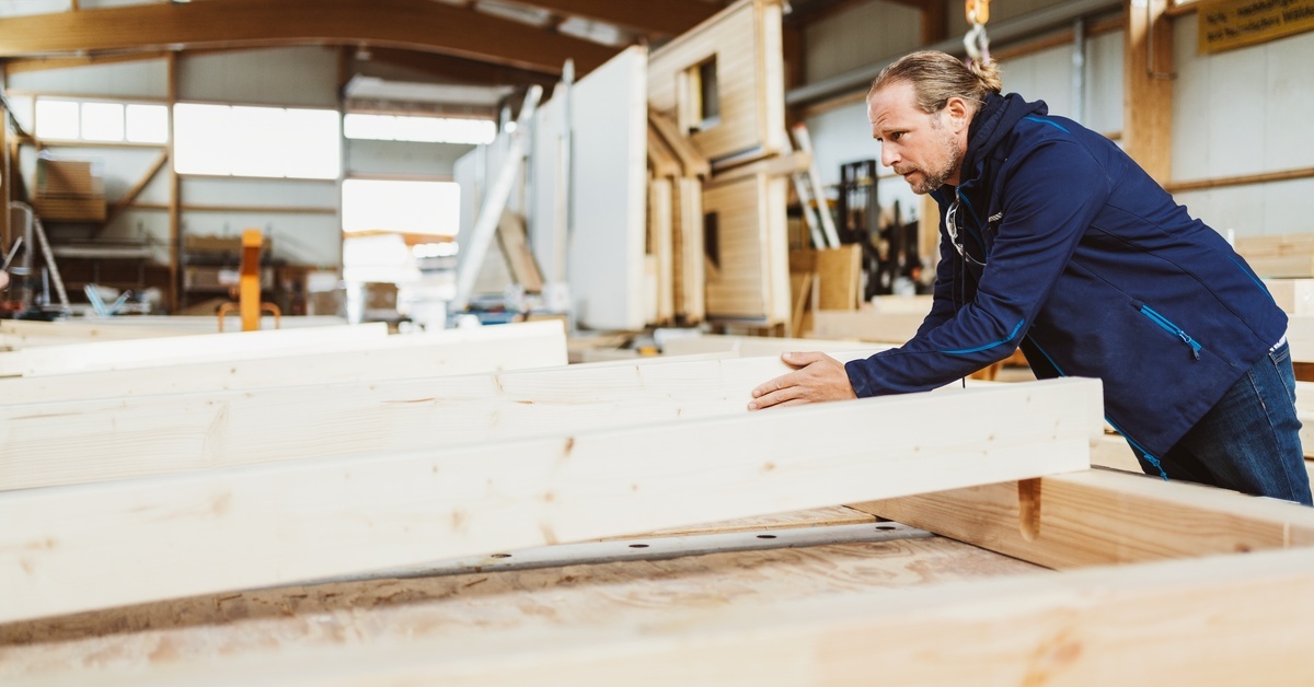 A carpenter slides a wooden support board into place while working on the framing of a prefabricated house.