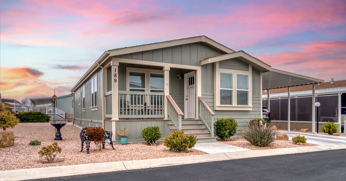 A craftsman-style gray manufactured home sits on a paved street in a neighborhood. Pink clouds color the sky at sunset.