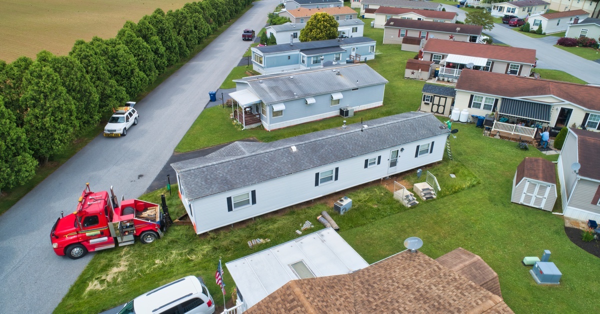 A aerial view of a red truck delivering a white manufactured home. The home sits in a mobile home park.