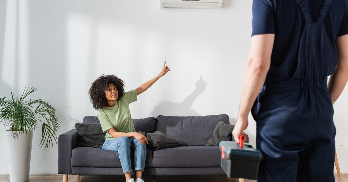 A young woman sits on a gray sofa in her living room and points at the AC unit while talking to the repair man.