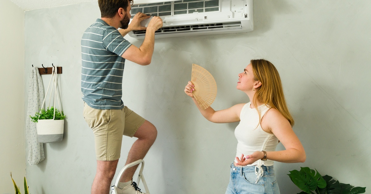A young couple tries to fix the split system AC unit in their living room. He stands on a ladder while she holds a fan.