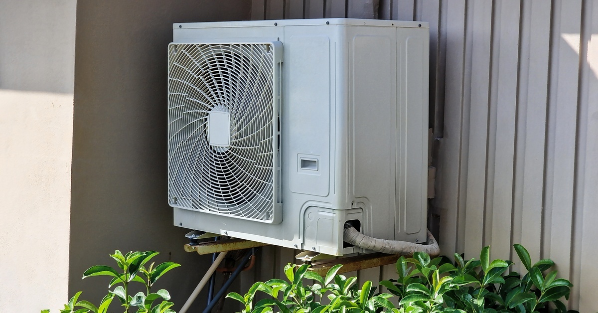 The outdoor condenser unit of an air conditioning system sits on brackets outside of a home. A green bush sits below it.