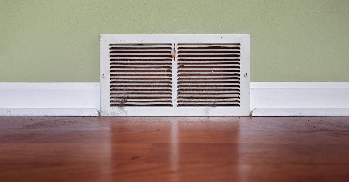 A dusty wall vent is installed along the baseboard of a home with a hardwood floor and a light green wall.