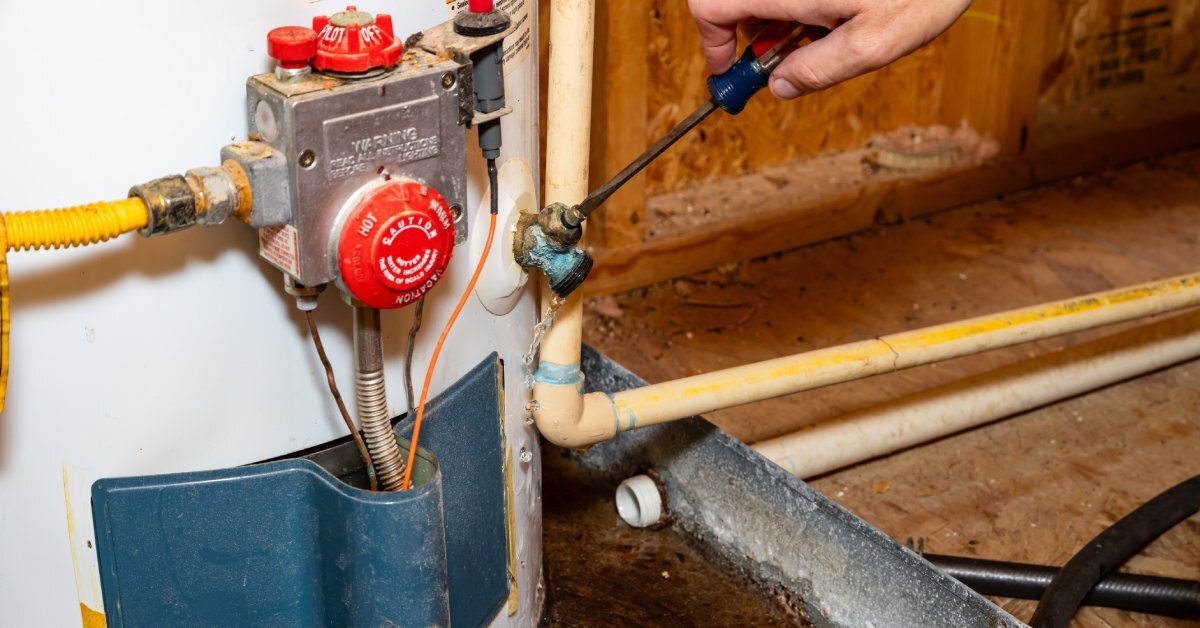 A person uses a screwdriver to close the drain valve on a gas water heater. Water drips from the drain valve.
