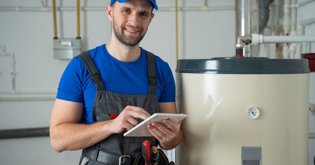 A smiling service technician stands next to a residential water heater while using a digital tablet.