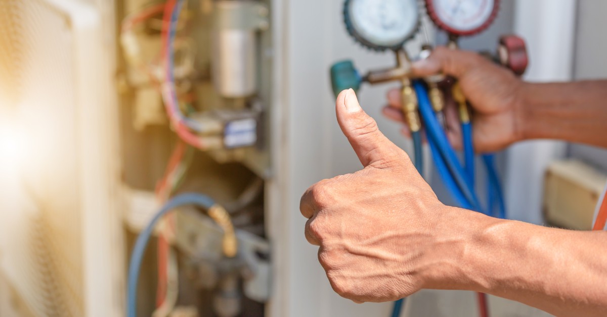 A HVAC technician's hand gives a thumbs-up to the camera. The technician is using tools to fix the AC unit.