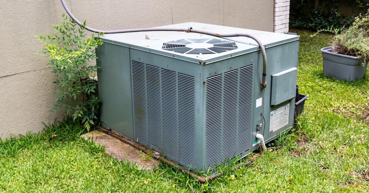 A packaged air conditioning unit is installed next to a tan home. The home has a grassy yard and a potted plant.