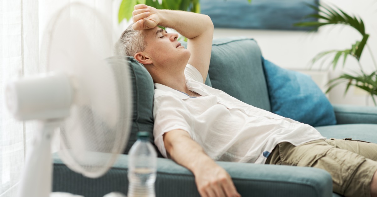 A tired man relaxes on his sofa while trying to stay cool. A fan sits on a nearby table next to bottled water.
