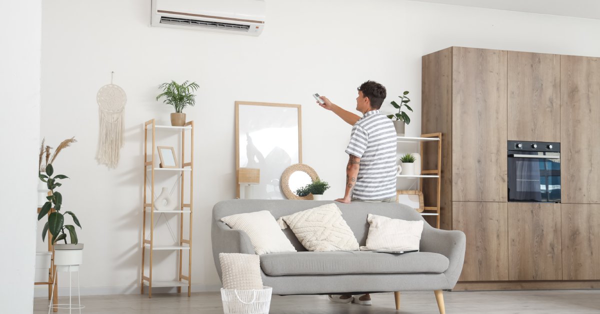 A young man stands in his living room with his back to the camera. He points a remote at the split AC unit on the wall.