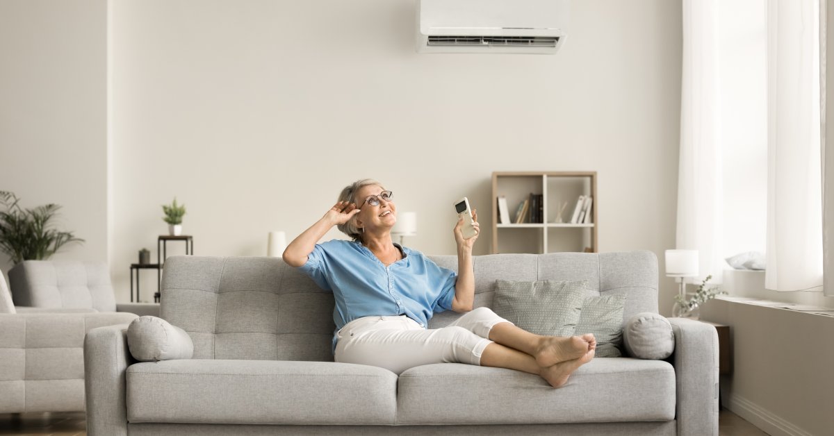 A smiling senior woman relaxes on her sofa with her shoes off. She holds the remote for the split AC system.
