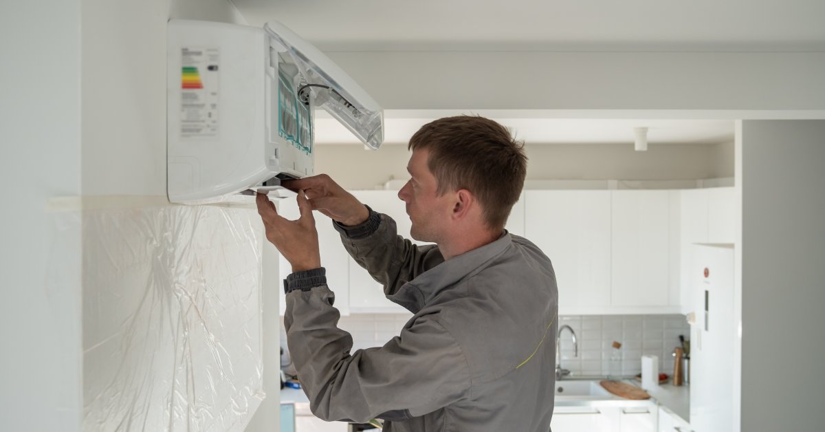 A technician installs the indoor blower for a split AC system on the wall near a kitchen with white cabinets and tile.