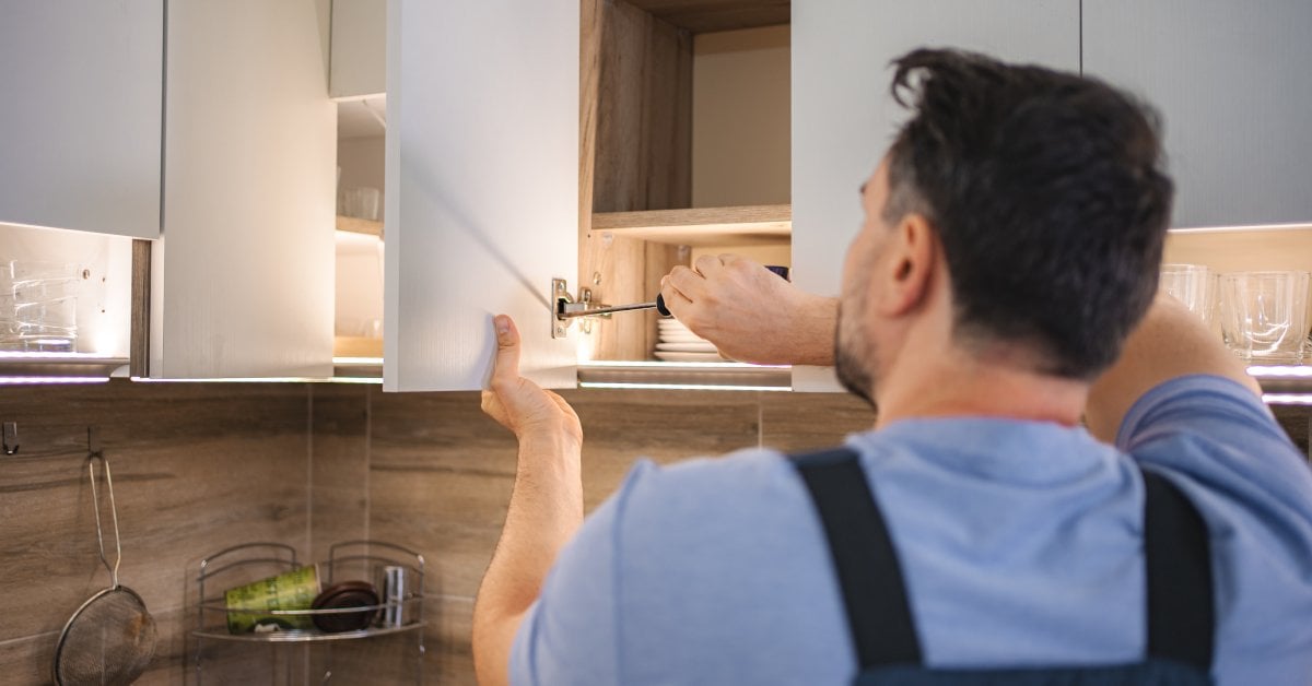 A man uses a screwdriver to tighten the hinge on a white kitchen cabinet. A wire shelf sits on the kitchen counter.