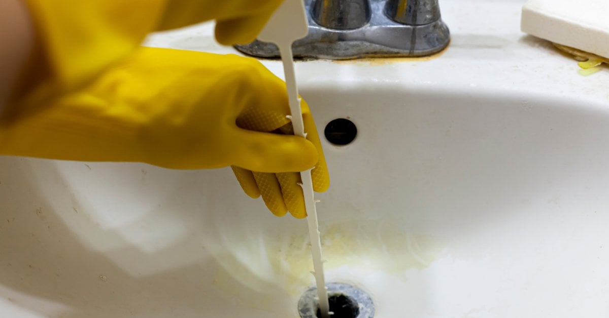 A person wearing yellow rubber gloves uses a white plastic drain snake to unclog a bathroom sink drain.
