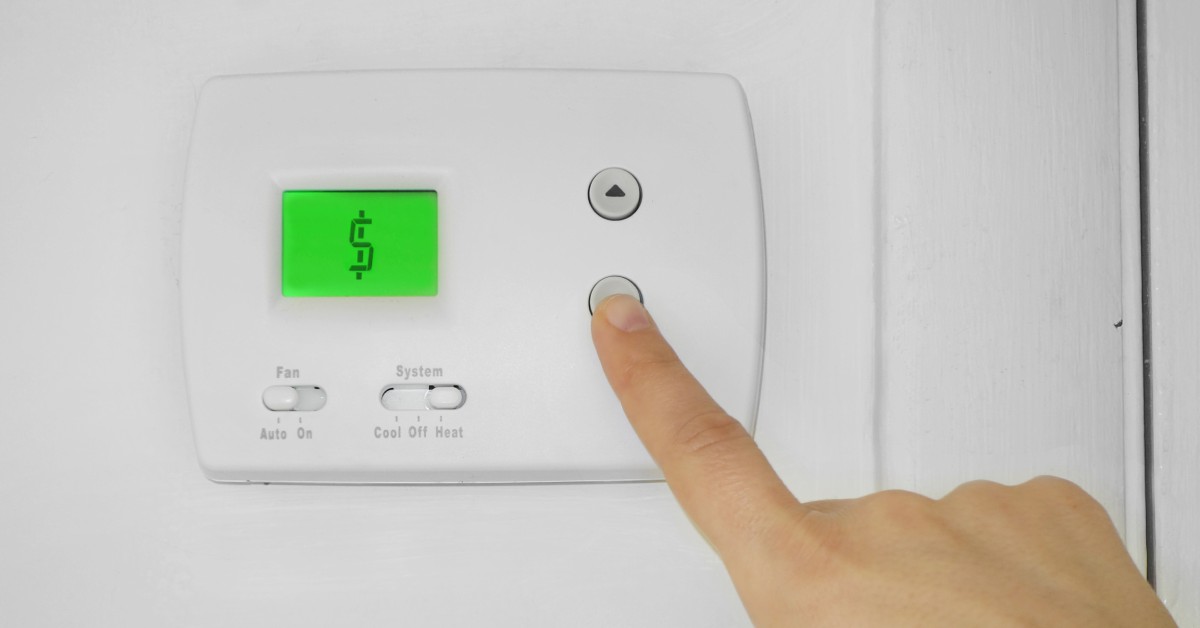 A finger presses the down arrow on a programmable thermostat. The green digital screen displays a dollar sign.