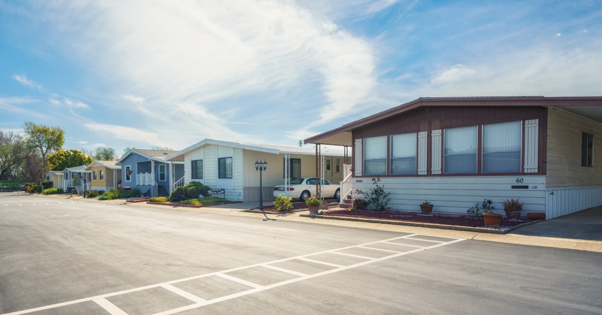 The sun shines on a row of modular homes that line a wide blacktop street. A crosswalk is painted on the street.